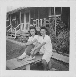 [Two young women sitting on wooden platform in front of barracks, 1-4-A and 1-4-B, Rohwer, Arkansas, October 1, 1944]