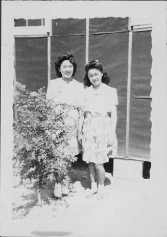 [Two young women standing in front of barracks wall next to shrub, Rohwer, Arkansas]
