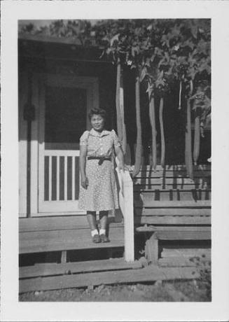 [Woman in patterned dress standing on barracks porch steps next to hechima gourds, Rohwer, Arkansas]