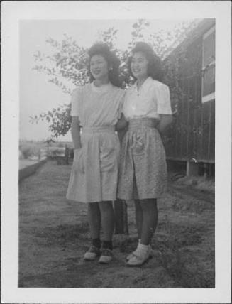 [Two young women standing side-by-side in front of small tree and barracks, full-length portrait, Rohwer, Arkansas]
