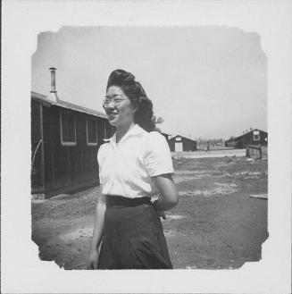 [Woman with dimples and eyeglasses standing in open area between barracks, three-quarter portrait, Rohwer, Arkansas]